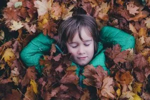 Child is Well-Settled in the New Environment. Child in green shirt lying in pile of brown maple leaves. Annie Spratt (Unsplash).
